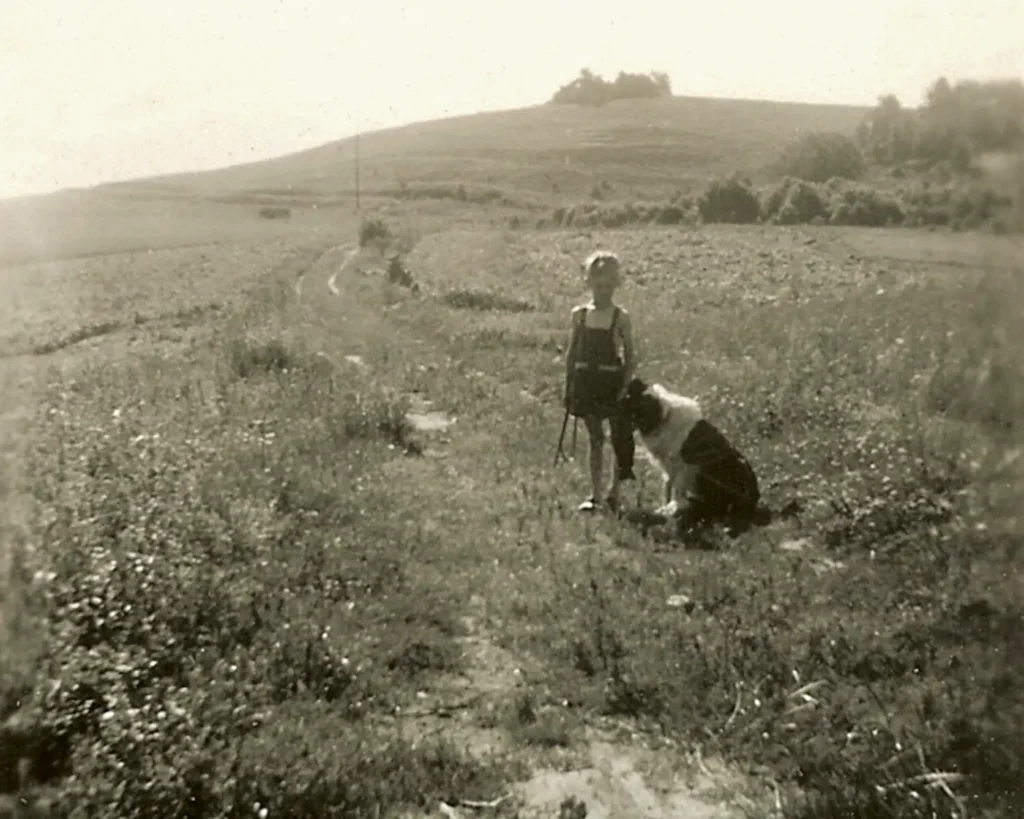 En liten pojke med hund står på en igenvixen väg med en backe bakom. Svartvitt foto.