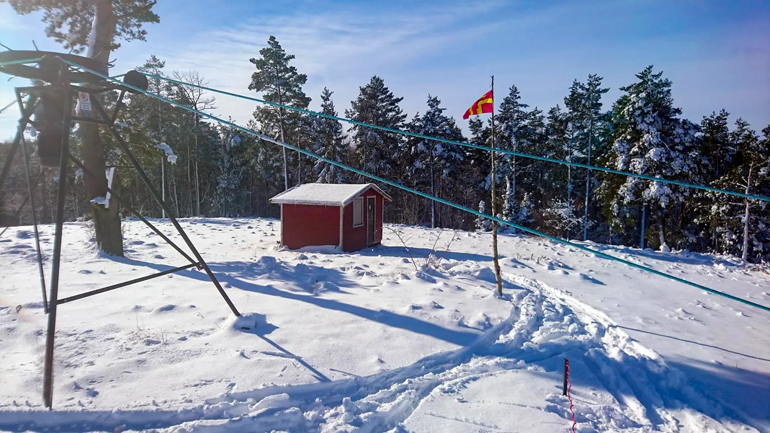 Övre vändhjulet på en skidlift med en röd toppstuga i fonden. Skånsk rödgul flagga vajar i vinden på en flaggstång av björk.
