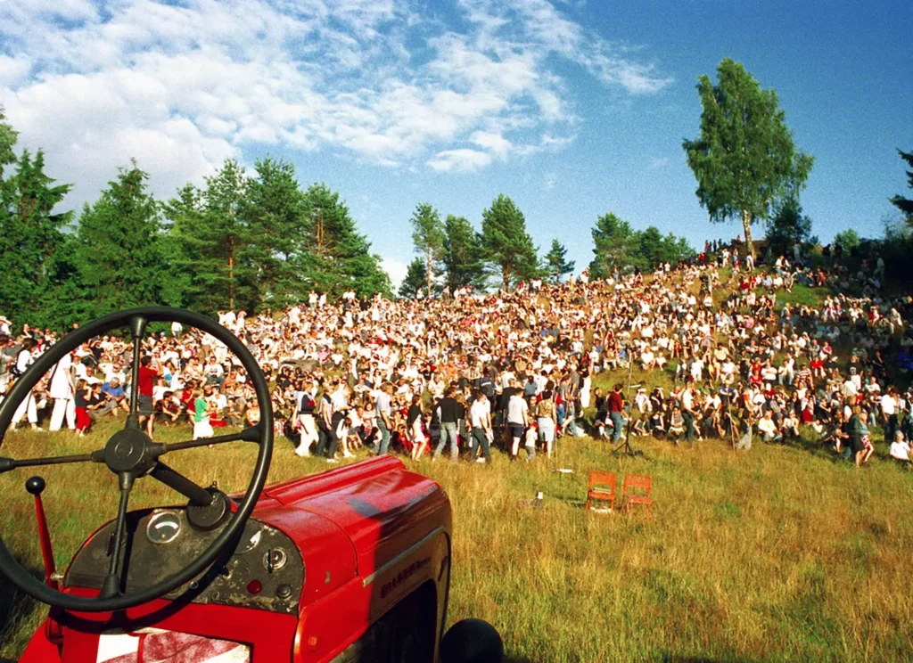 Tusentals människor sitter i en skidbacke som en amfiteater för att titta på en traktorkonsert i botten av backen. I förgrunden en röd traktor.
